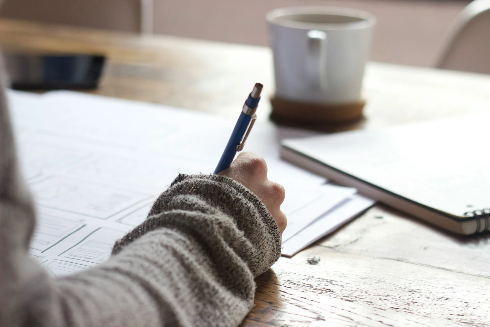 Person in a gray sweater sketches on paper with a blue pencil at a wooden desk beside a white coffee mug and tablet.