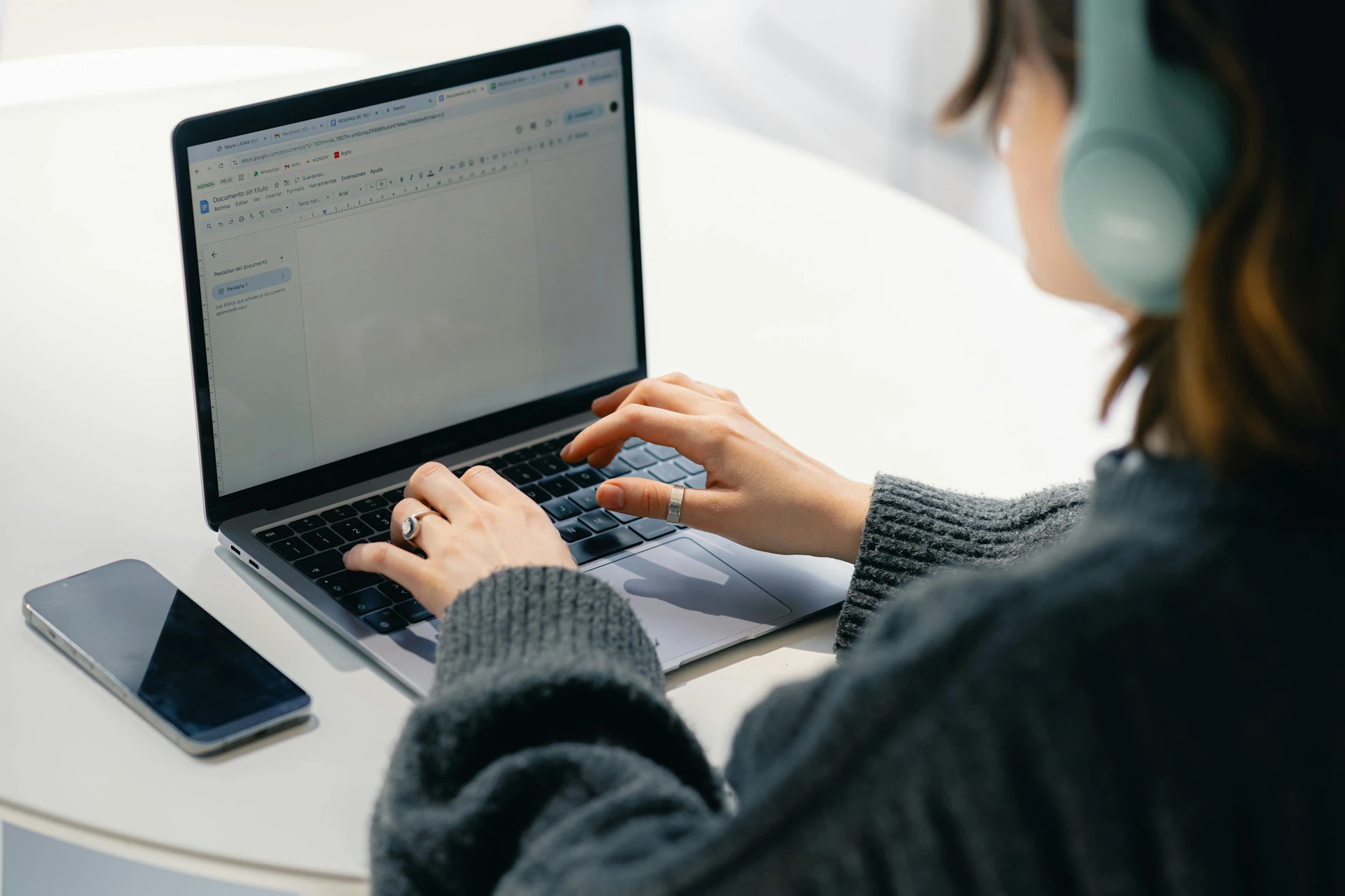 Woman typing on a laptop displaying a document at a white desk, with a smartphone beside her.