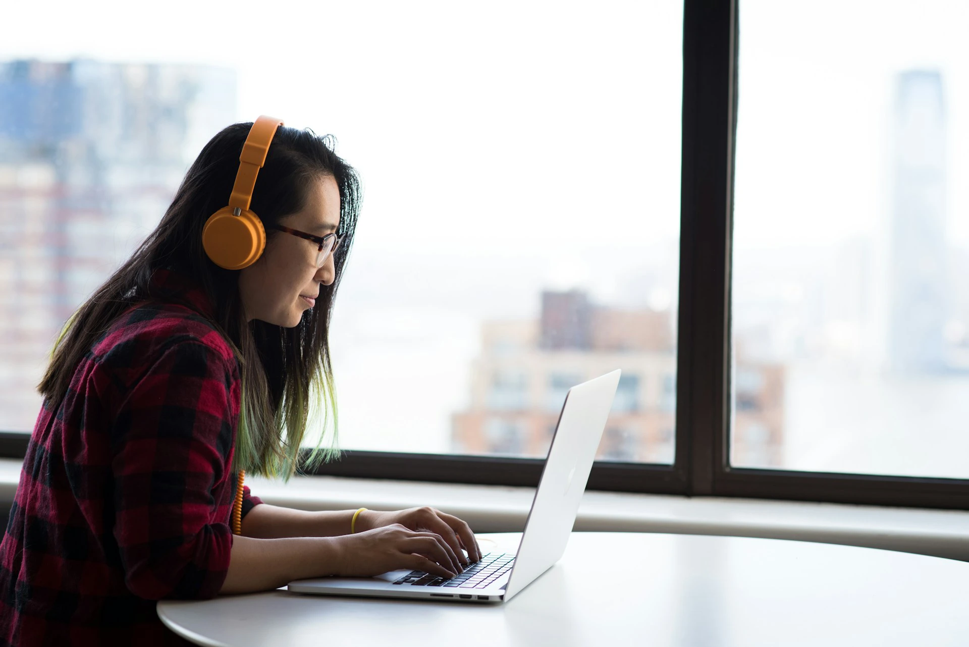 Woman wearing orange headphones and glasses works on a laptop at a table beside a large window overlooking city buildings.