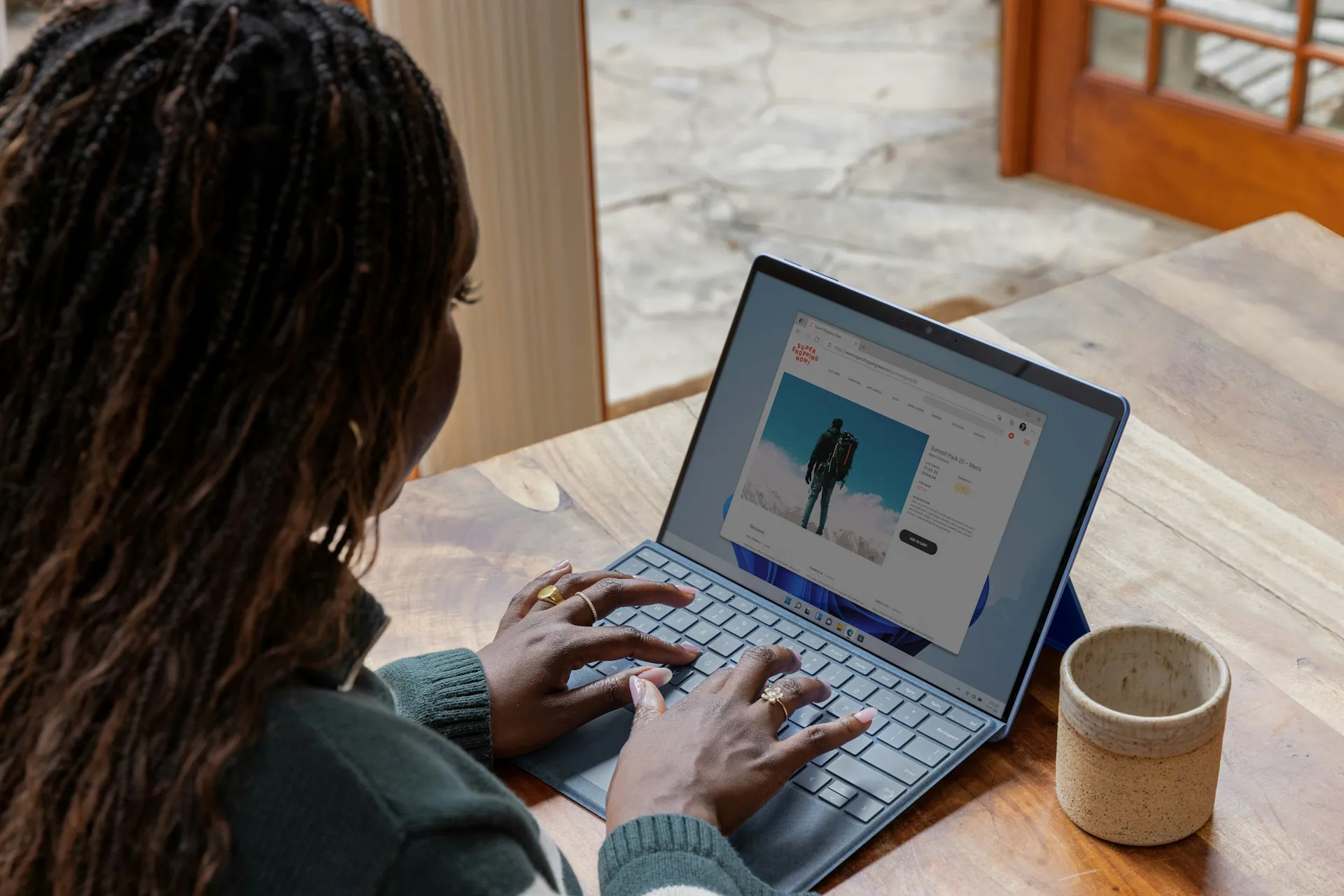Person with long braids typing on a laptop at a wooden table beside a cup of coffee, viewing a webpage with a product image.