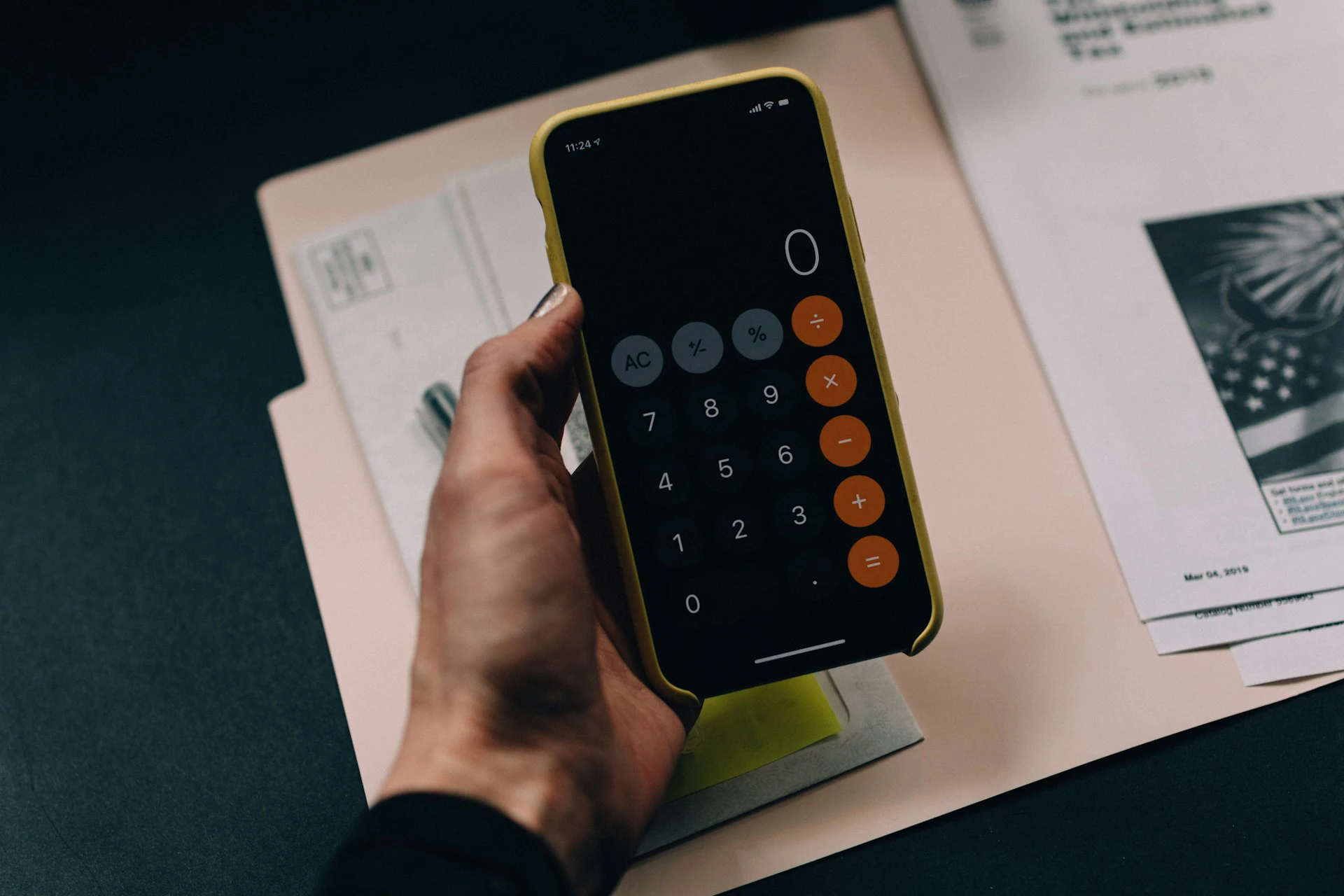 Hand holding a smartphone displaying a calculator app over paperwork, a folder, and a printed document on a desk.