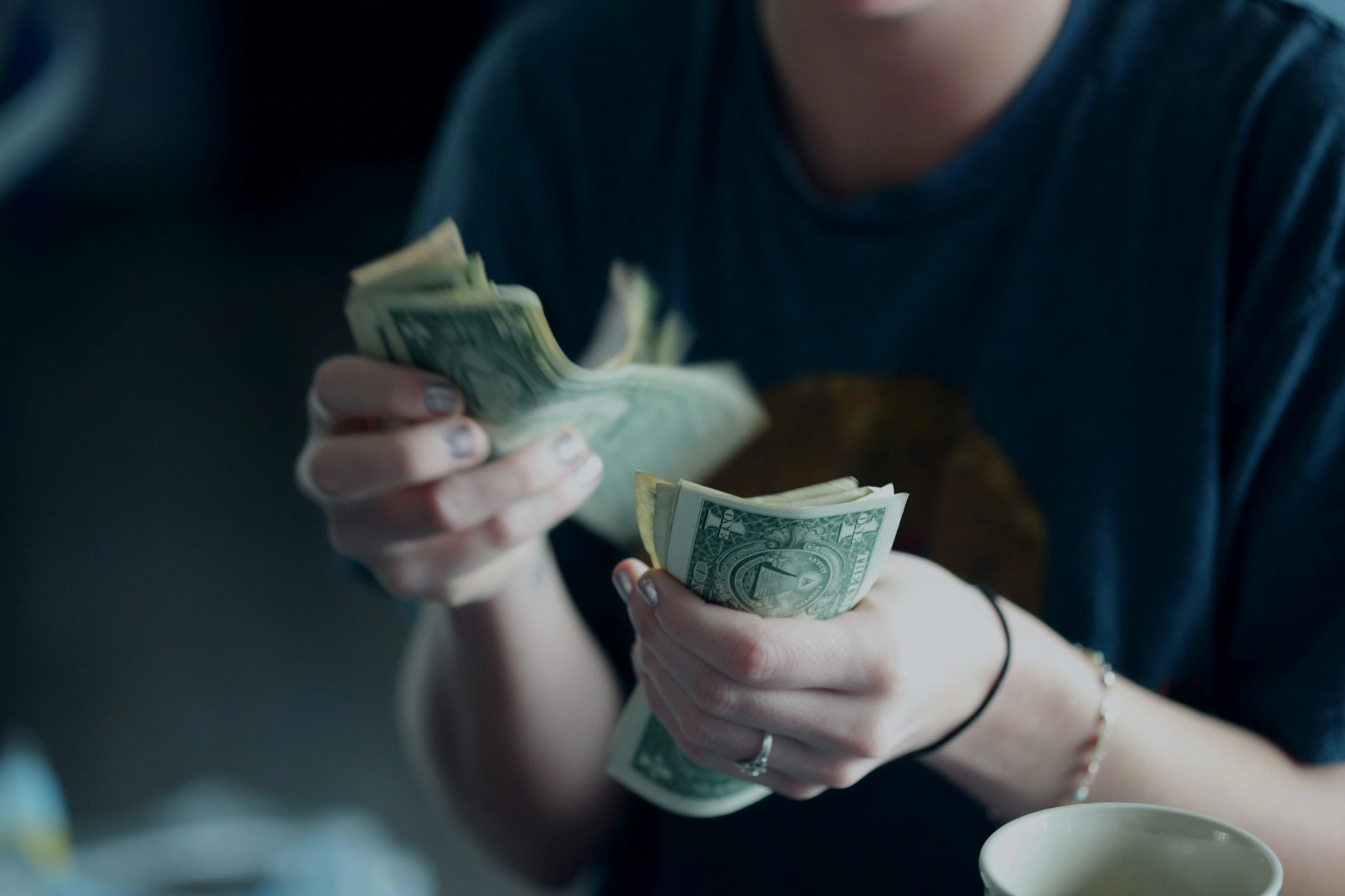 Person counting stacks of U.S. dollar bills beside a coffee cup.