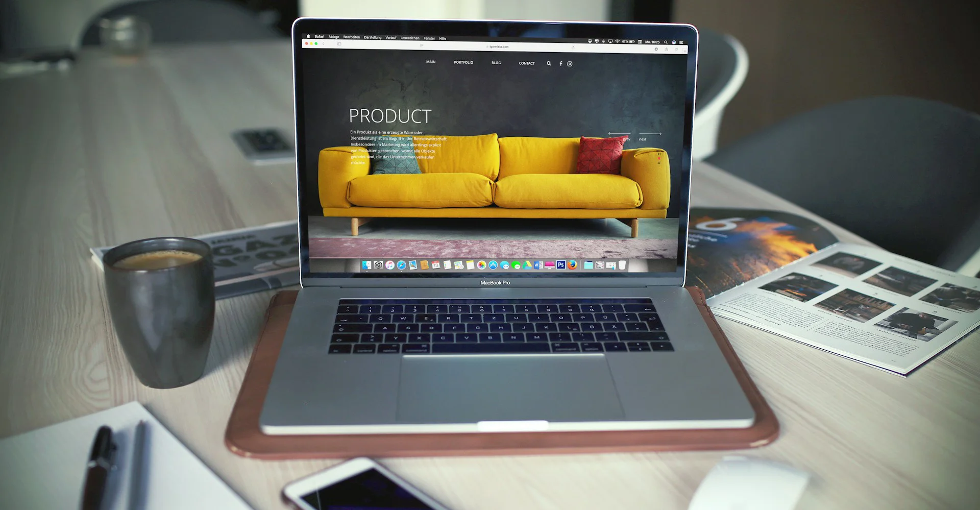 Laptop displaying a furniture website with a yellow sofa on a desk beside coffee, magazines, and a smartphone.