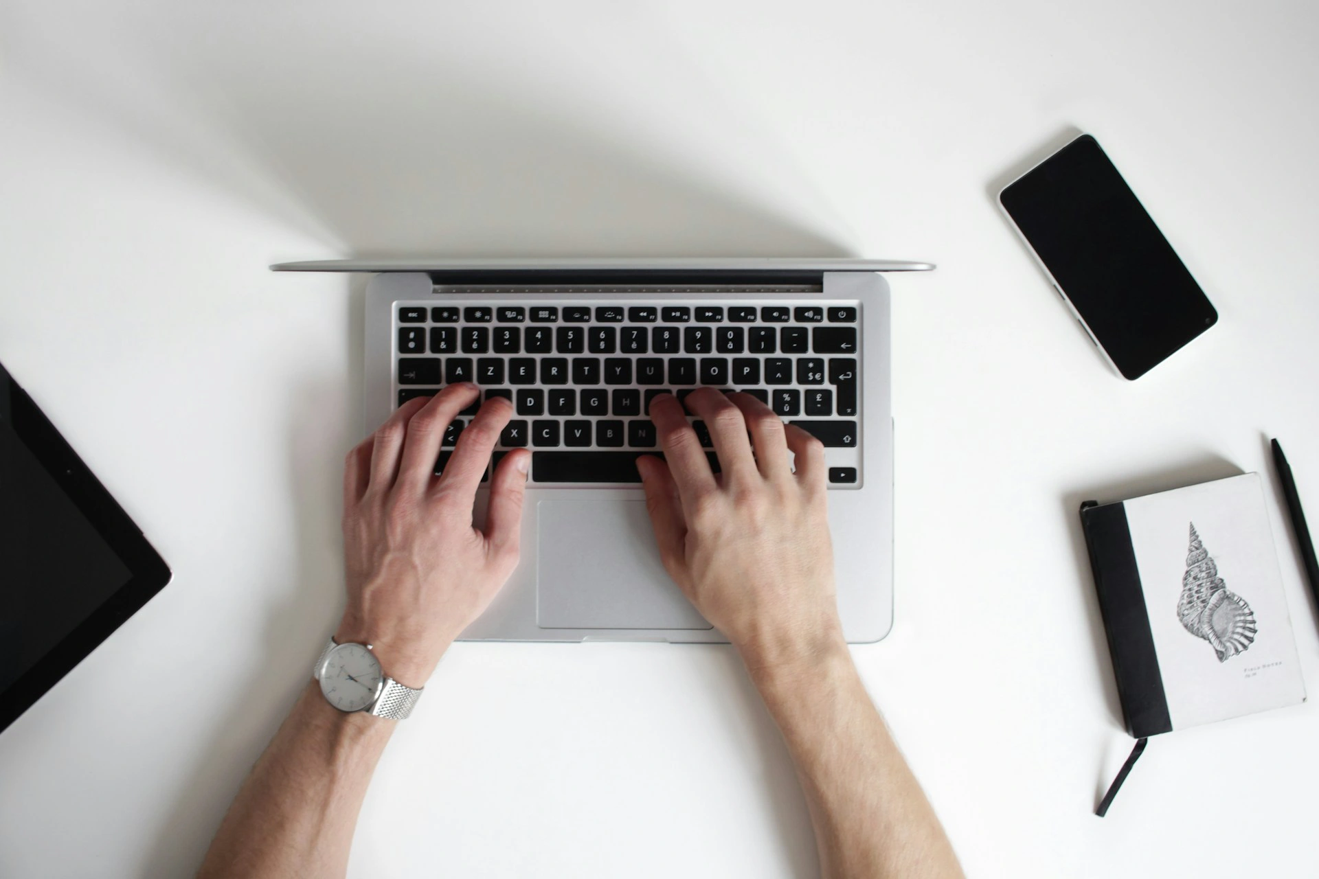 Hands typing on a laptop at a white desk with a smartphone, tablet, notebook, and pen nearby.