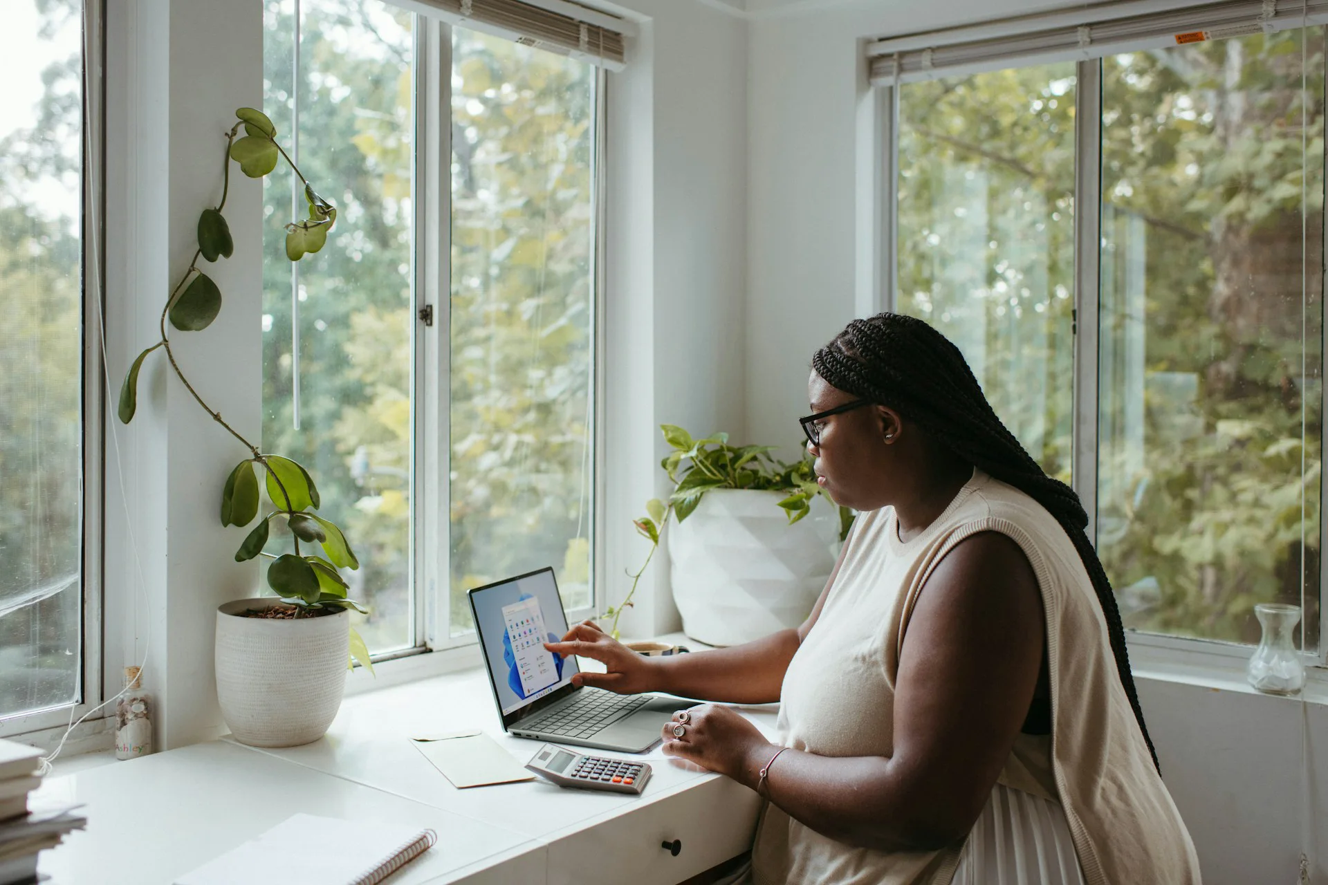 Woman working on a laptop at a bright window-side desk, with a calculator, notebook, and potted plants nearby.