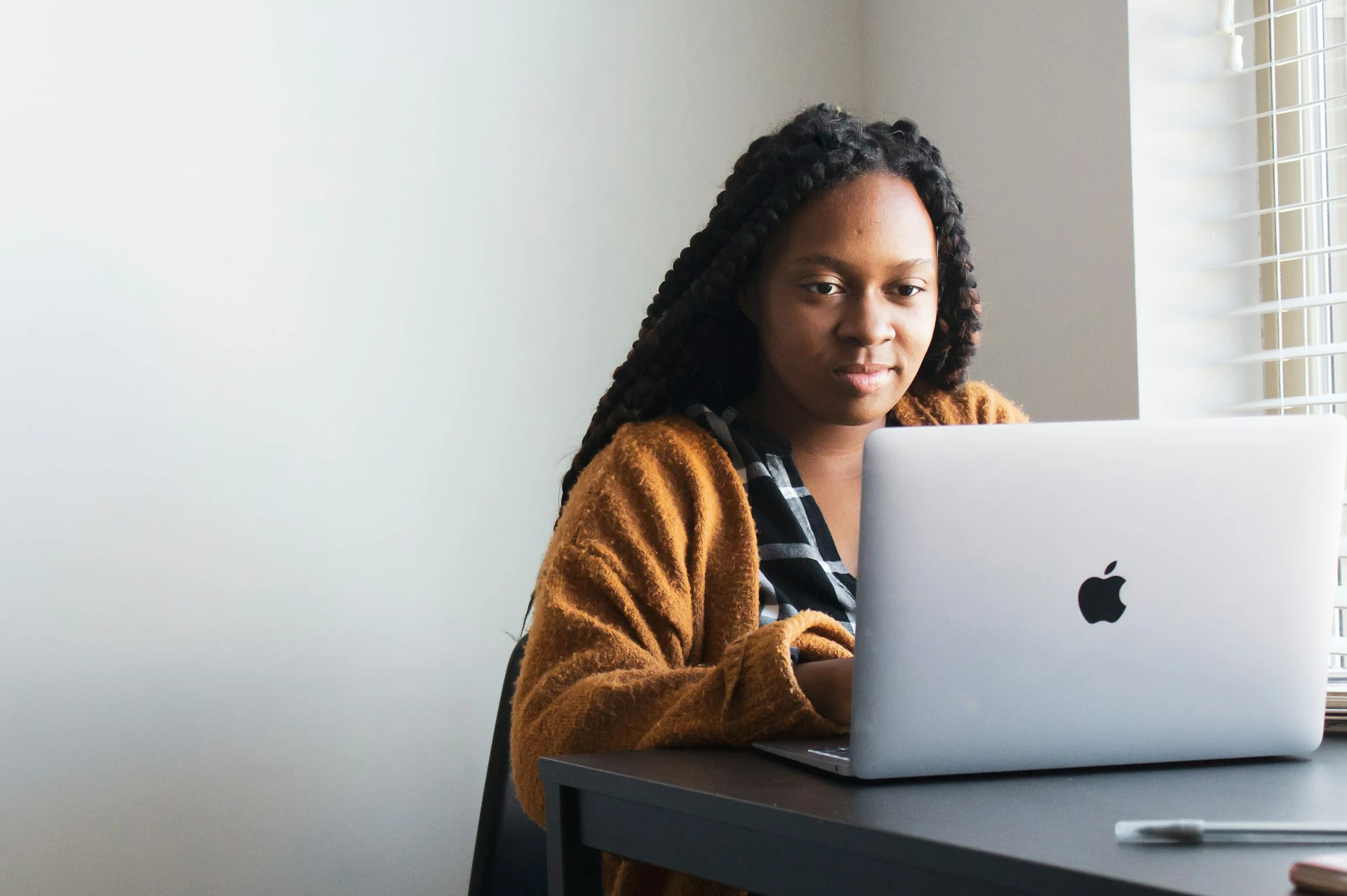 A woman with long braids works on an Apple laptop at a table beside a sunlit window.