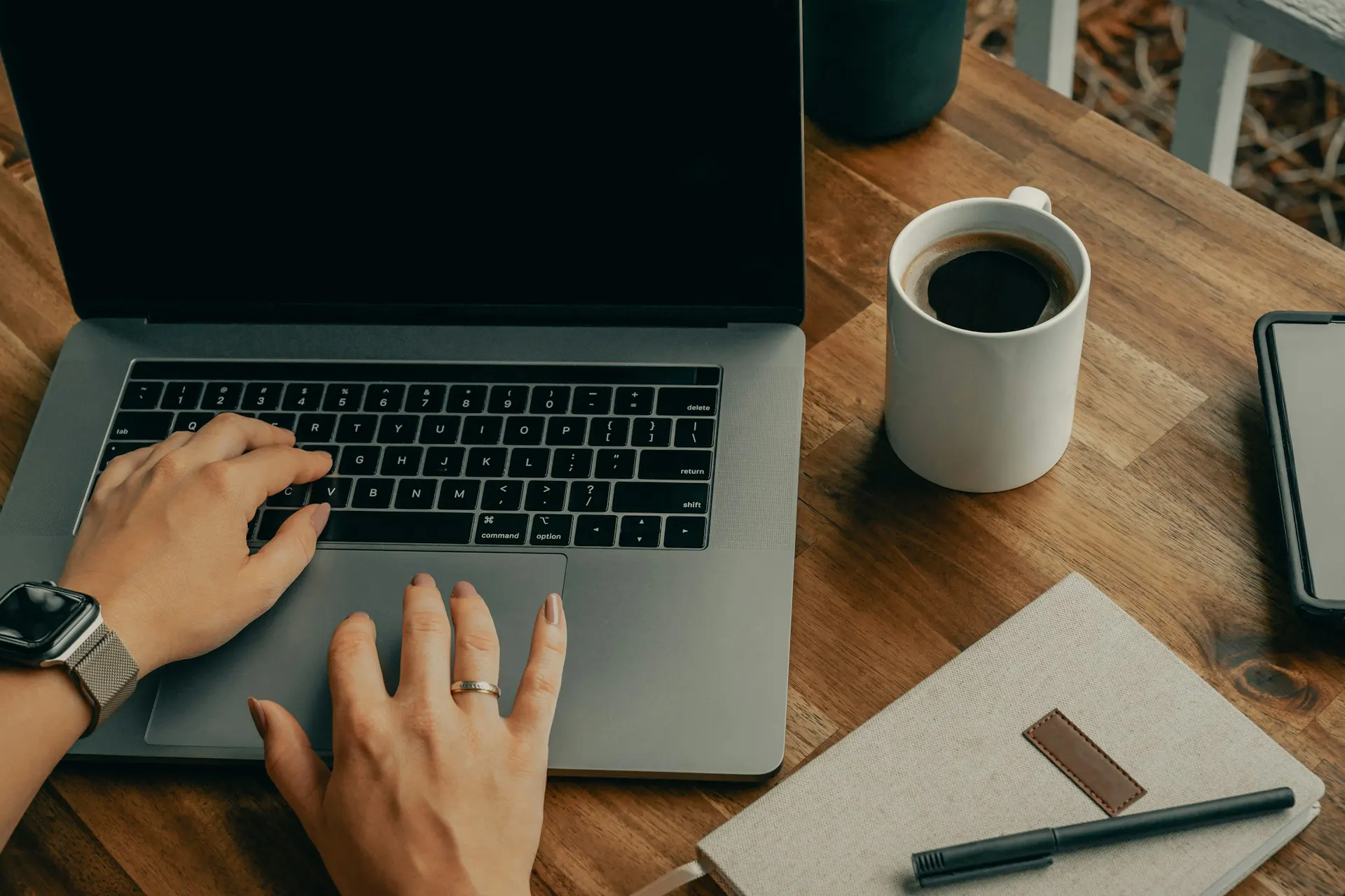 A person wearing a smartwatch types on a laptop at a wooden desk, beside a white mug, tablet, notebook, and pen.