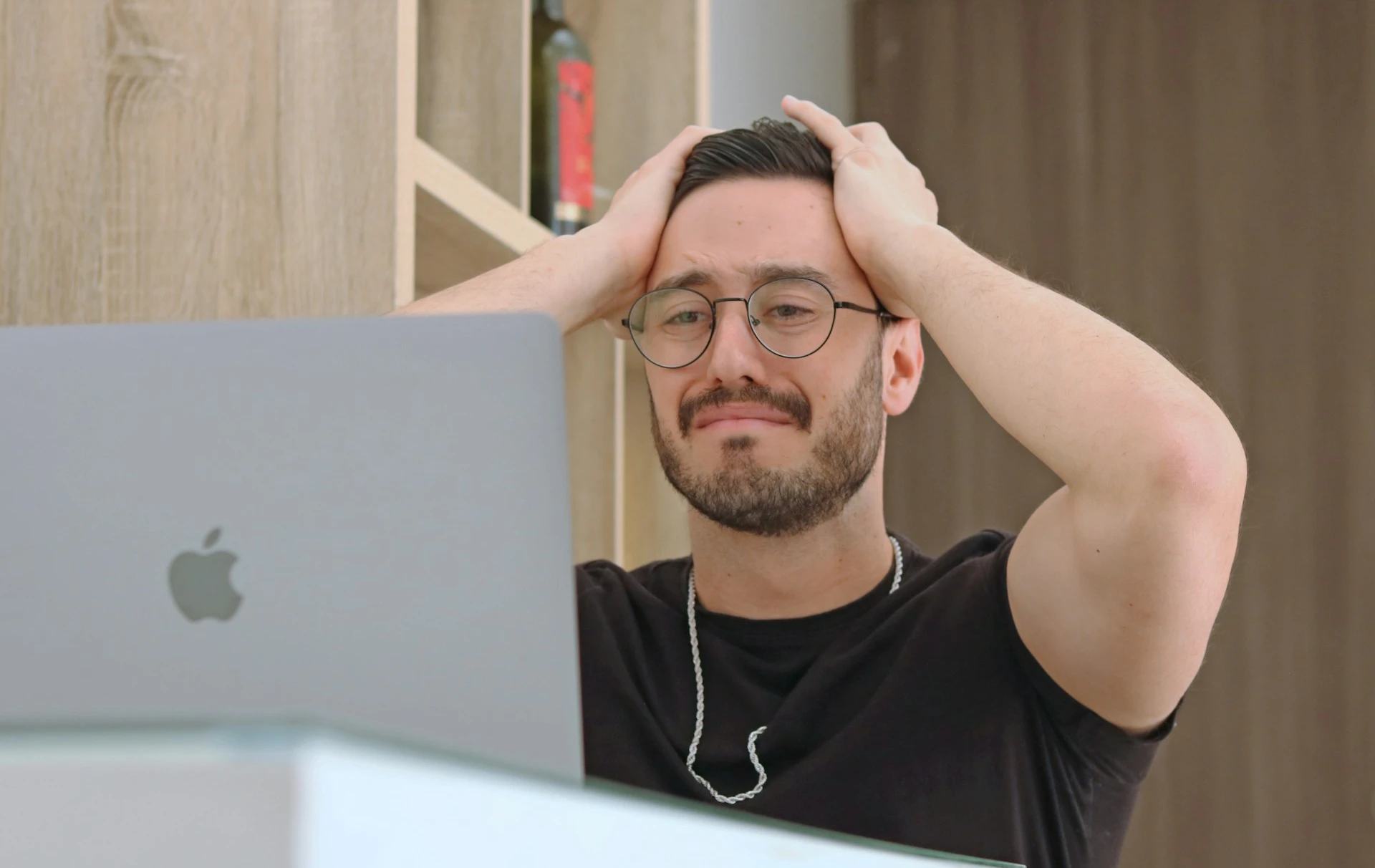 A man wearing round glasses sits behind an open Apple laptop, holding both hands on his head as he looks at the screen.