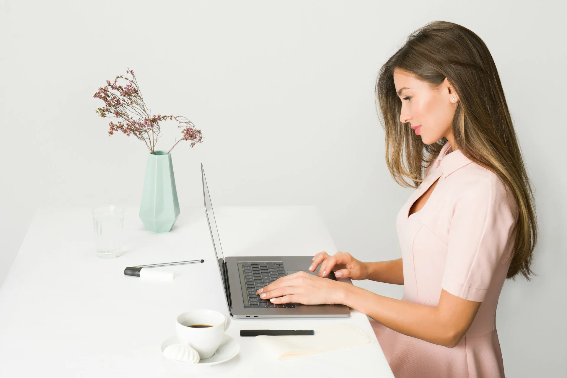 A woman in a pink blouse types on a laptop at a white table with coffee, a pen, and a vase of flowers.
