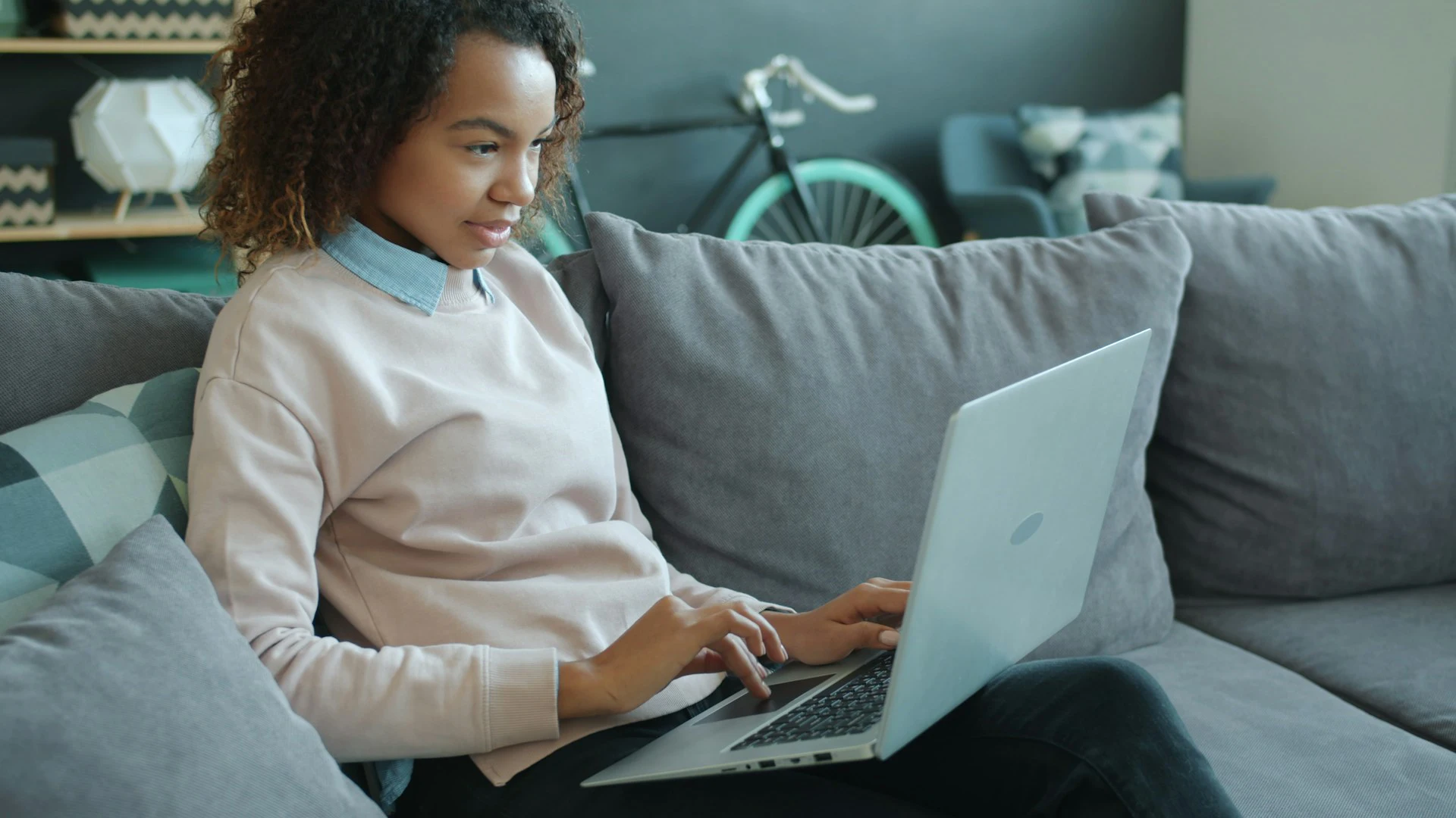 A woman in a white sweater sits on a gray sofa typing on a silver laptop, with a bicycle visible in the room behind her.