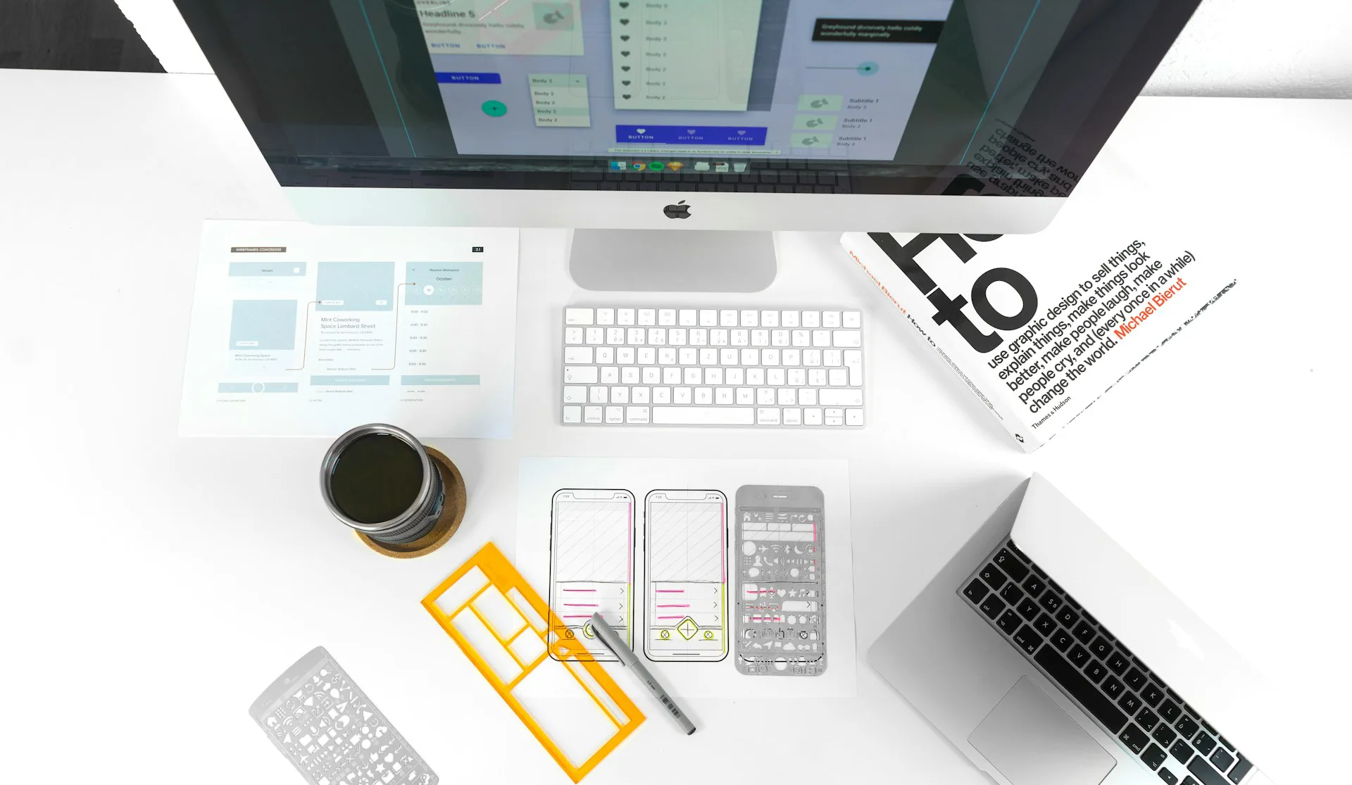 Top-down workspace with an iMac, keyboard, laptop, coffee, design sketches, smartphone wireframes, a ruler, and a design book.
