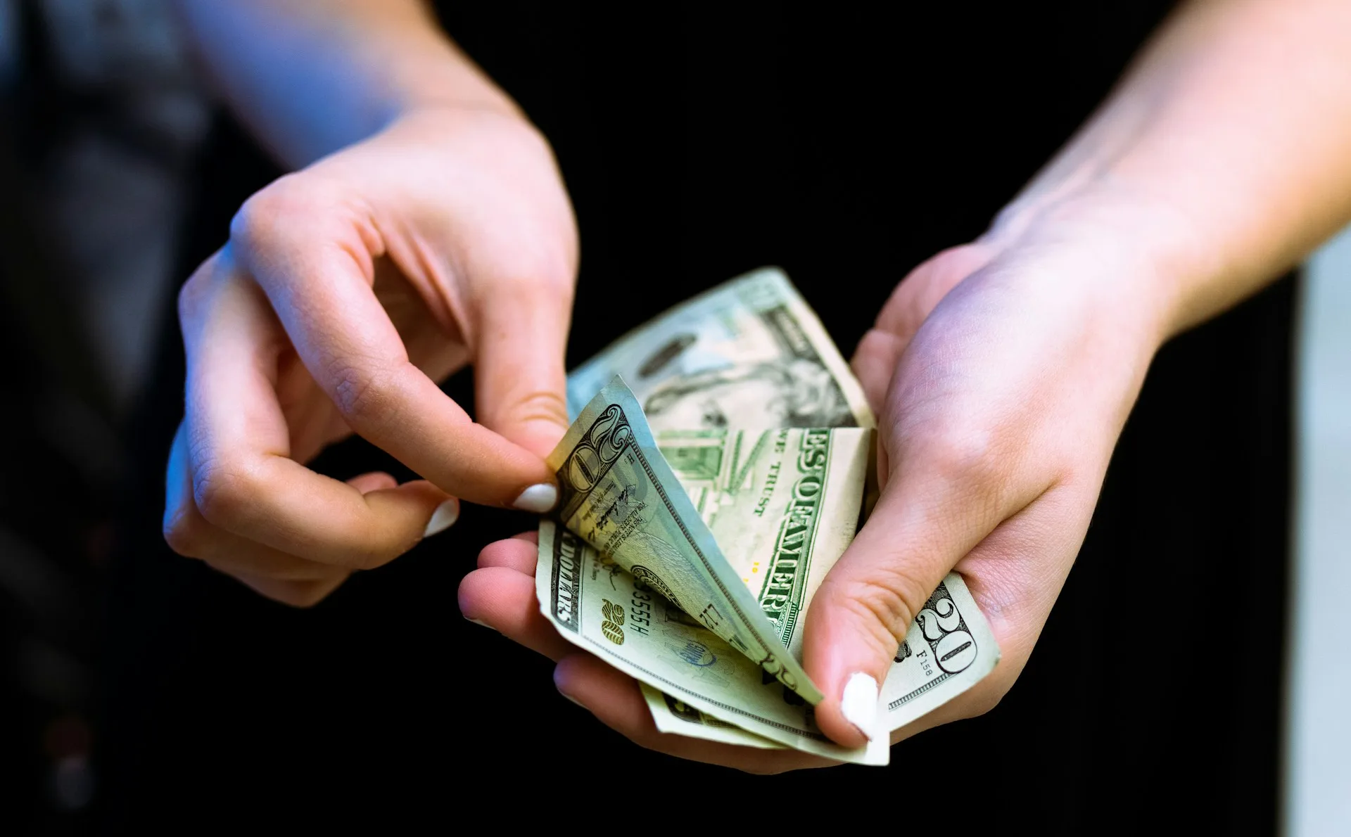 Two people’s hands count a stack of U.S. dollar bills, including visible $20 and $50 notes, against a dark background.